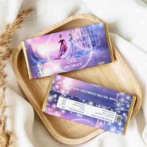 Pu&ograve; includere: Due barrette di cioccolato con involucro in lamina d'oro sono esposte su un vassoio di legno. La barretta superiore presenta un design viola e rosa con la scritta "Amelia's 5th BIRTHDAY!" e immagini di personaggi animati. La barretta inferiore ha un'etichetta bianca.