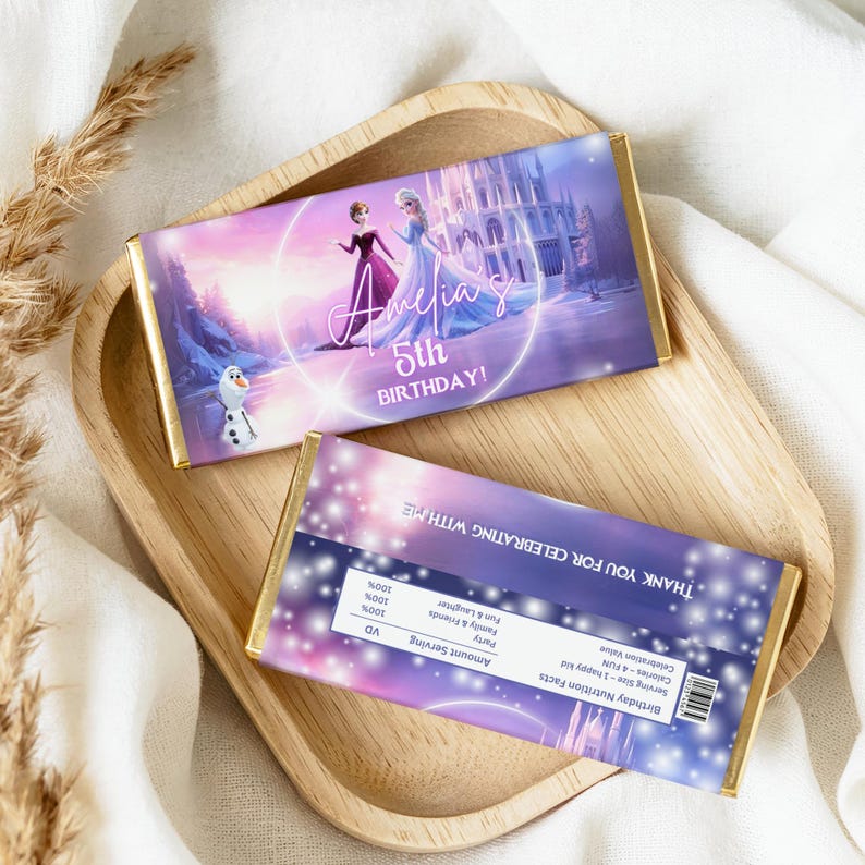 Pu&ograve; includere: Due barrette di cioccolato con involucro in lamina d'oro sono esposte su un vassoio di legno. La barretta superiore presenta un design viola e rosa con la scritta "Amelia's 5th BIRTHDAY!" e immagini di personaggi animati. La barretta inferiore ha un'etichetta bianca.