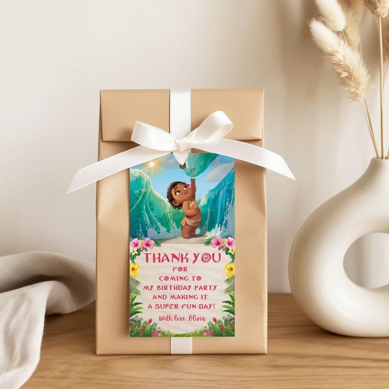 A gift wrapped with a blue ribbon and decorated with a baby Moana thank you tag. The tag stands out with vibrant ocean colors, tropical flowers, and a cute island baby character.