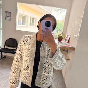 Peut inclure: Cardigan en crochet fait à la main, sans fermeture, avec un motif de carrés granny. La veste présente une combinaison de carrés blancs et gris, avec une bordure et des manches grises unies. Les manches sont longues et le cardigan arrive aux genoux.