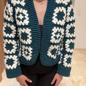 Peut inclure: Un cardigan en crochet bleu canard et blanc. Le pull sans fermeture présente un motif de carrés granny avec une base bleu canard et des motifs floraux blancs. Les manches et le col sont bleu canard uni.