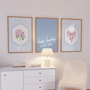 Puede incluir: Tres impresiones artísticas enmarcadas con fondos a rayas azules y blancas. Una impresión presenta una hortensia rosa, otra tiene el texto "how lucky are we", y la tercera muestra un lazo rosa. Los marcos son de color marrón claro.