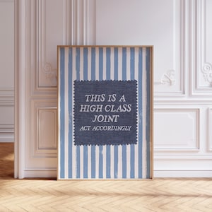 May include: A framed print with a blue and white striped design. The print has a dark blue square with the text "THIS IS A HIGH CLASS JOINT ACT ACCORDINGLY" in white. The frame is a light brown colour.