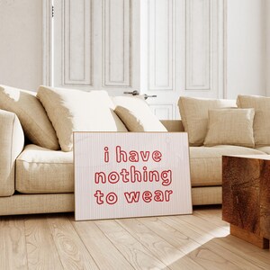 Framed wall art print featuring the phrase “i have nothing to wear” in bold red bubble letters against a white background with thin, vertical red wavy lines. The minimalist and playful design is illuminated by soft natural light, creating a cozy and modern bedroom aesthetic.