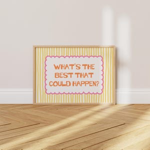 May include: A rectangular framed print featuring a white central panel with a pink scalloped border and the orange text "WHAT'S THE BEST THAT COULD HAPPEN?" set against a background of vertical yellow and white stripes within a light wood frame.