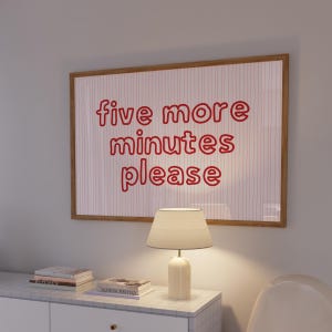 Puede incluir: Un cuadro enmarcado con las palabras "five more minutes please" en letras rojas. El cuadro está montado en una pared encima de un mueble blanco con una lámpara y libros.