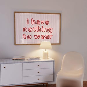 Framed wall art print featuring the phrase “i have nothing to wear” in bold red bubble letters against a white background with thin, vertical red wavy lines. The minimalist and playful design is illuminated by soft natural light, creating a cozy and modern bedroom aesthetic.
