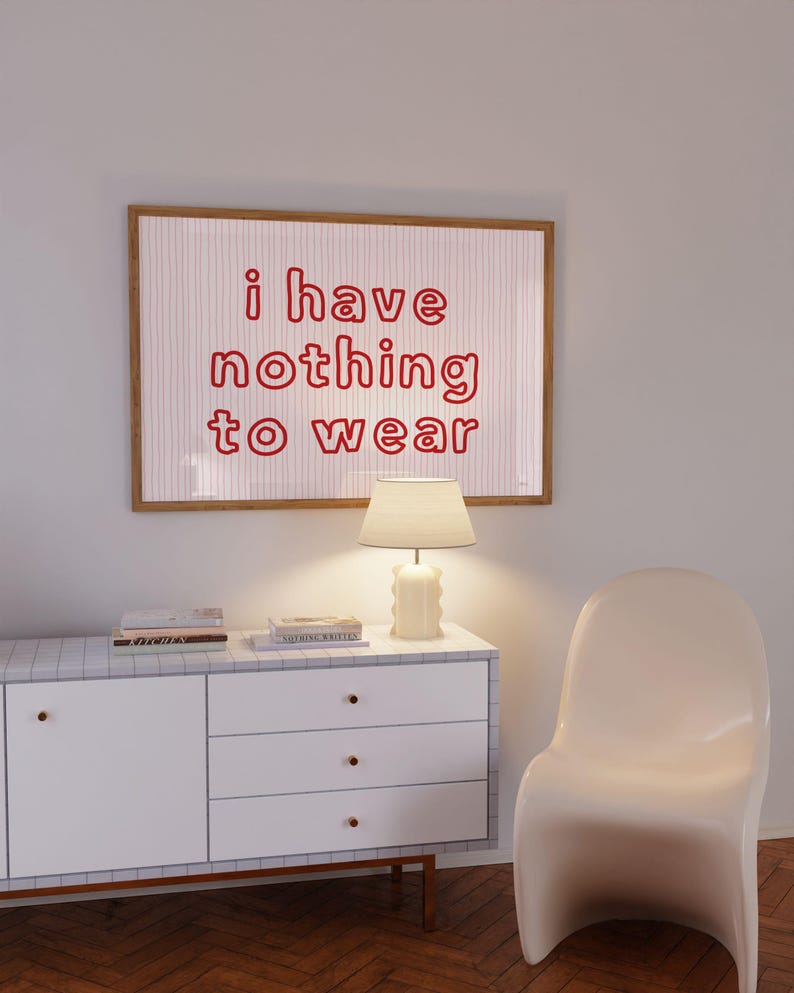 Framed wall art print featuring the phrase “i have nothing to wear” in bold red bubble letters against a white background with thin, vertical red wavy lines. The minimalist and playful design is illuminated by soft natural light, creating a cozy and modern bedroom aesthetic.