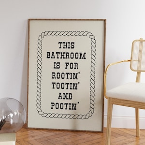May include: A rectangular framed wall art piece with a light beige background and a black rope-style border. The text in black reads: THIS BATHROOM IS FOR ROOTIN' TOOTIN' AND POOTIN'. The frame is made of a medium-toned brown wood material.