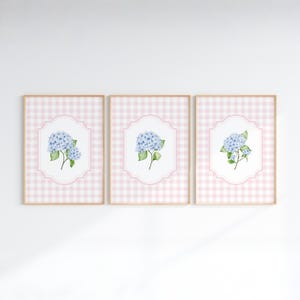 May include: A set of three framed wall art prints featuring light blue hydrangea flower illustrations centered within white scalloped frames, all set against a soft pink and white gingham checkered background pattern in thin wooden frames.