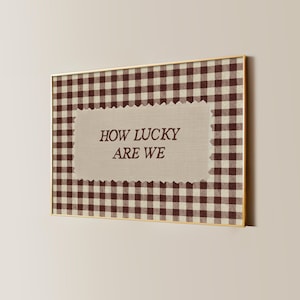 Könnte beinhalten: Ein gerahmtes Kunstwerk mit goldenem Rand. Das Kunstwerk zeigt ein braun-weißes Vichy-Muster, das ein beiges Rechteck mit dem Text "HOW LUCKY ARE WE" in braunen Buchstaben umgibt.