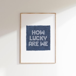 May include: Framed artwork featuring a square of blue fabric with the embroidered text "HOW LUCKY ARE WE" in white. The artwork is set against a white background, with a simple wooden frame.