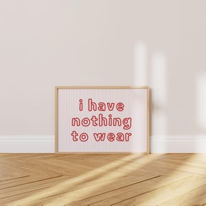 Framed wall art print featuring the phrase “i have nothing to wear” in bold red bubble letters against a white background with thin, vertical red wavy lines. The minimalist and playful design is illuminated by soft natural light, creating a cozy and modern bedroom aesthetic.