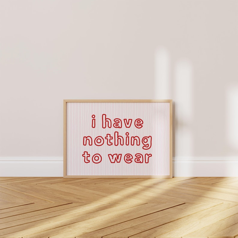 Framed wall art print featuring the phrase “i have nothing to wear” in bold red bubble letters against a white background with thin, vertical red wavy lines. The minimalist and playful design is illuminated by soft natural light, creating a cozy and modern bedroom aesthetic.