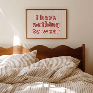 Framed wall art print featuring the phrase “i have nothing to wear” in bold red bubble letters against a white background with thin, vertical red wavy lines. The minimalist and playful design is illuminated by soft natural light, creating a cozy and modern bedroom aesthetic.