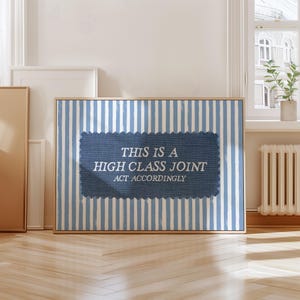 Op de afbeelding: Een ingelijst kunstwerk met een blauw-wit gestreepte achtergrond. Het kunstwerk heeft een denim patch met de tekst "THIS IS A HIGH CLASS JOINT ACT ACCORDINGLY" in het wit. De lijst is lichtbruin.