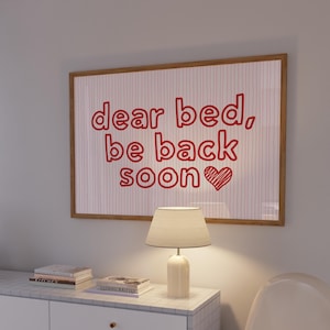Puede incluir: Un cuadro enmarcado con el texto "dear bed, be back soon" en rojo, acompañado de un corazón. La obra de arte cuelga sobre una lámpara blanca y una pequeña mesa con libros.