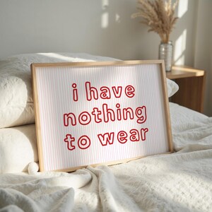 Framed wall art print featuring the phrase “i have nothing to wear” in bold red bubble letters against a white background with thin, vertical red wavy lines. The minimalist and playful design is illuminated by soft natural light, creating a cozy and modern bedroom aesthetic.