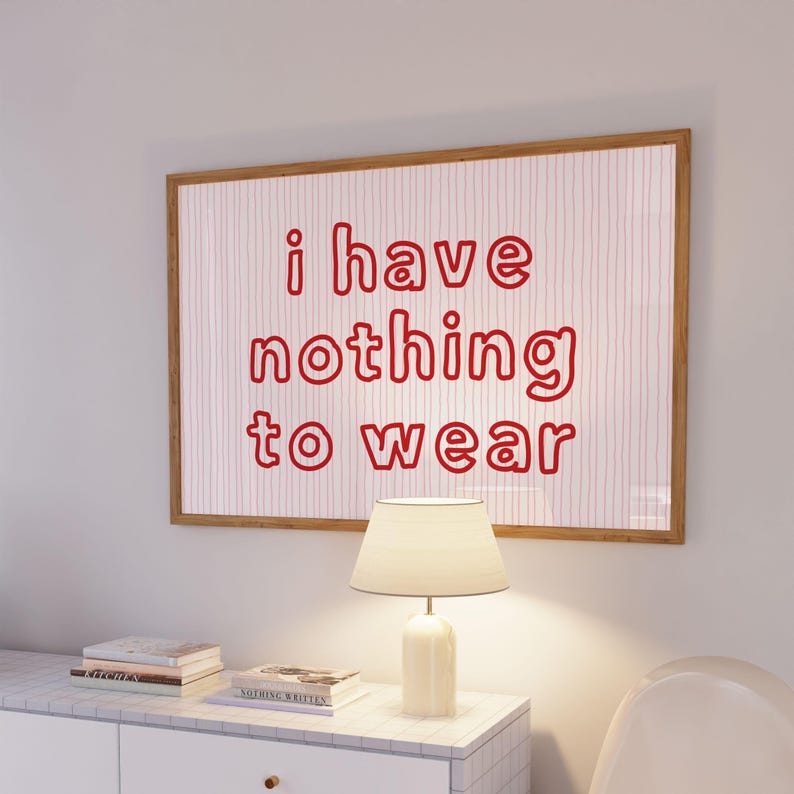 Framed wall art print featuring the phrase “i have nothing to wear” in bold red bubble letters against a white background with thin, vertical red wavy lines. The minimalist and playful design is illuminated by soft natural light, creating a cozy and modern bedroom aesthetic.
