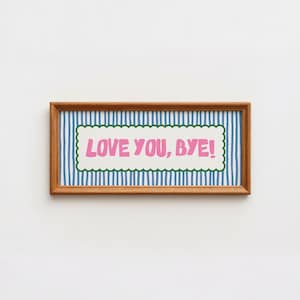 May include: A rectangular wooden frame containing a print with a blue and white vertical striped background. Centered is a cream-colored scalloped rectangle featuring the pink text "LOVE YOU, BYE!" in a bold, rounded, sans-serif typeface.
