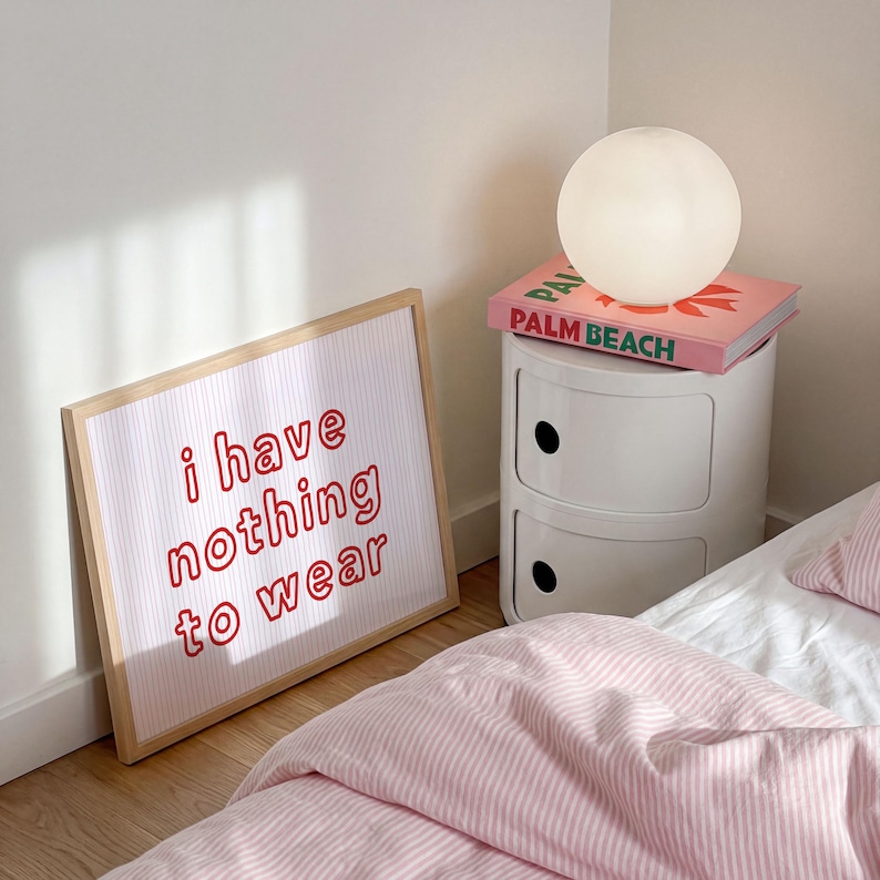 Framed wall art print featuring the phrase “i have nothing to wear” in bold red bubble letters against a white background with thin, vertical red wavy lines. The minimalist and playful design is illuminated by soft natural light, creating a cozy and modern bedroom aesthetic.