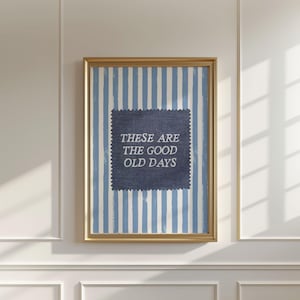 May include: A framed wall art print featuring a vertical light blue and white striped background. Centered is a dark blue square patch with pinked edges containing the white text: THESE ARE THE GOOD OLD DAYS. The frame is a metallic gold colour.