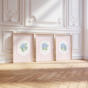 May include: Three rectangular framed wall art prints featuring a light pink and white gingham pattern background. Each print displays a central white scalloped label containing a watercolor illustration of a blue hydrangea flower with green leaves.