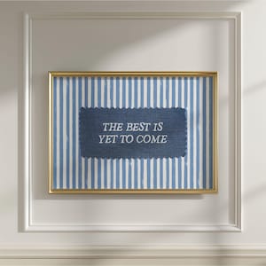 Puede incluir: Una pieza de arte mural rectangular en un marco dorado con un fondo de rayas verticales azules y blancas. Centrado hay un parche texturizado azul oscuro con bordes festoneados que muestra el texto en blanco, THE BEST IS YET TO COME.