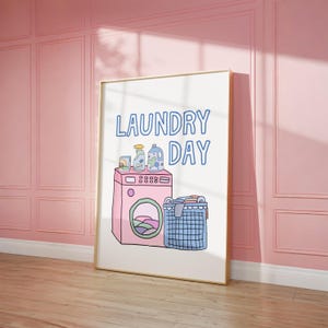 Könnte beinhalten: Ein gerahmter Druck mit den Worten "LAUNDRY DAY" in blauer Schrift. Die Illustration zeigt eine rosa Waschmaschine, Reinigungsmittel und einen blauen Wäschekorb voller Kleidung. Das Kunstwerk ist vor einem weißen Hintergrund.