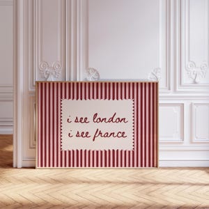 May include: A rectangular framed wall art piece featuring a vertical dark red and light pink striped background. Centered on the stripes is a cream-colored rectangular label with scalloped edges containing the text i see london i see france.
