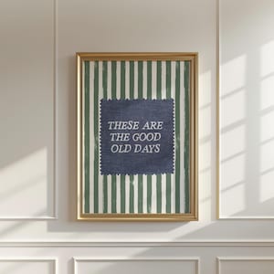 Pode incluir: Arte de parede emoldurada com uma moldura dourada. A obra de arte apresenta um quadrado azul com o texto "THESE ARE THE GOOD OLD DAYS" em um fundo listrado verde e branco. A arte está pendurada em uma parede branca.