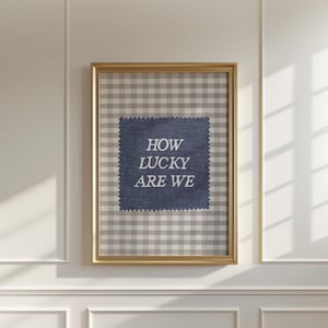 May include: Framed wall art with a gold-colored frame. The artwork features a gray and white gingham pattern background. A navy blue square with the text "HOW LUCKY ARE WE" is centered on the artwork.