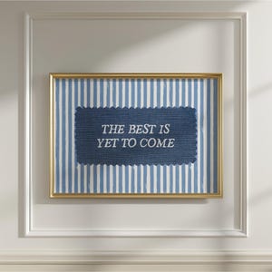 以下が含まれることがあります： 金色のフレームに入ったプリント。青と白のストライプの背景に、中央に濃い青色の長方形のパッチがあります。「THE BEST IS YET TO COME」というテキストがパッチに表示されています。
