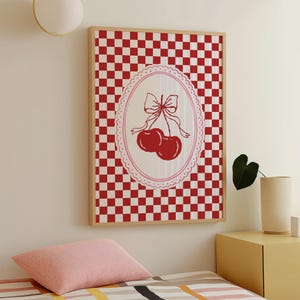 May include: Framed wall art with a red and white chequered background. The artwork features a white oval with a red cherry illustration and a bow. The frame is made of light-coloured wood.