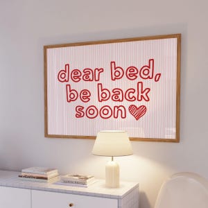 May include: A rectangular wall art print in a light wood frame featuring the red text "dear bed, be back soon" with a hand-drawn red heart symbol. The background of the print consists of thin, vertical red stripes on a white surface.
