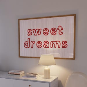 Könnte beinhalten: Ein gerahmtes Kunstwerk mit den Worten "sweet dreams" in roter Schrift hängt über einem weißen Schrank. Eine cremefarbene Lampe mit Lampenschirm steht auf dem Schrank, neben einem Stapel Bücher. Das Kunstwerk hat einen hellbraunen Rahmen.