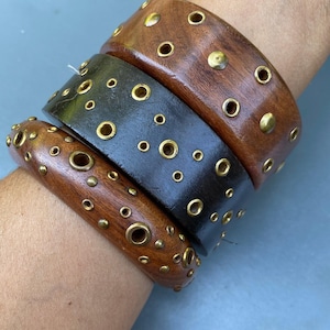 May include: Three wooden bangles with brass accents. The top and bottom bangles are a medium brown, while the middle one is black. Each bangle is studded with brass rivets and eyelets, creating a bold, textured look. These bracelets are a statement piece.