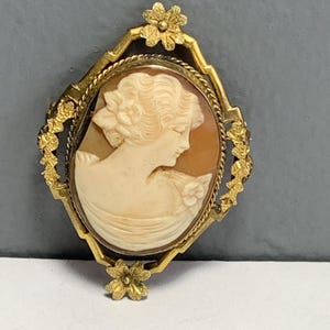 May include: A vintage cameo brooch featuring a profile of a woman carved in cream-colored shell. The oval cameo is set in a gold-toned frame with floral and rope detailing. The brooch has a pin closure on the back.