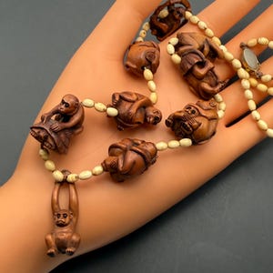 May include: A necklace featuring carved wooden monkey figures strung on a cream-colored beaded chain. The monkeys are in various poses, with detailed expressions and textures. The necklace is displayed on a hand.