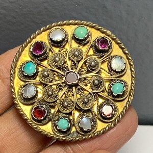 May include: A circular, gold-toned brooch with a filigree design. The brooch is decorated with turquoise, pink, and white stones.
