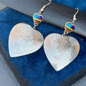 May include: A pair of silver heart-shaped earrings with intricate floral designs. Each earring features a smaller heart-shaped charm with colorful inlay.