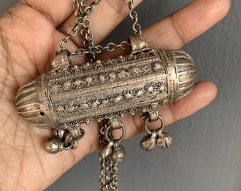 May include: A silver pendant necklace with a long, cylindrical charm that is intricately detailed with floral patterns. The charm has a small loop at the top and hangs from a chain with multiple strands. The pendant is adorned with small, dangling bells.