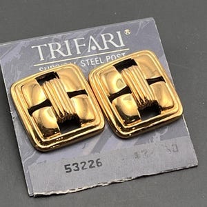 May include: A pair of gold-tone Trifari earrings with a geometric design. The earrings are square-shaped with a cutout in the center. The earrings are on a card with the brand name "Trifari" and the number "53226".