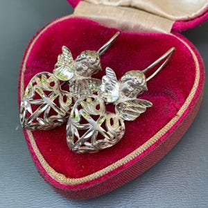May include: A pair of silver earrings with a heart-shaped design featuring an angel with wings. The earrings are displayed in a red velvet heart-shaped box.