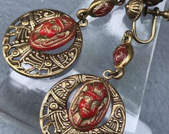 Puede incluir: Un par de pendientes colgantes de tono dorado con caras de esmalte rojo y diseños filigranados intrincados.