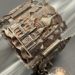 Puede incluir: Brazalete de metal plateado con un relieve detallado de la Basílica del Sagrado Corazón en París. El brazalete tiene un diseño calado con patrones intrincados y la palabra "PARIS" en un dije.