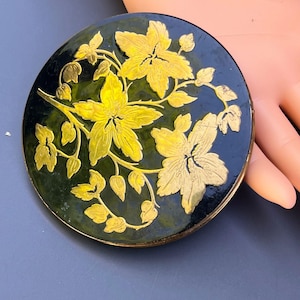 Op de afbeelding: Een ronde, zwart-groene compact met een gouden bloemenontwerp. De bloemen zijn gestileerd en hebben een reliëf.