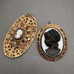 May include: Two gold-toned cameo pendants with black and white cameo portraits. One pendant has a filigree design with rhinestones. The other pendant has a simple oval frame.