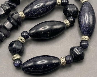 Puede incluir: Un collar negro y plateado con cuentas ovaladas y redondas. Las cuentas están hechas de una piedra azul oscuro con una textura moteada.