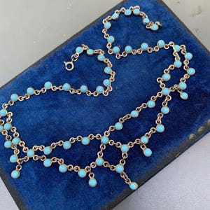 Op de afbeelding: Een delicate zilverkleurige ketting met turkooizen stenen. De ketting heeft een kettingontwerp met kleine, ronde turkooizen stenen langs de ketting en hangend aan de onderkant.